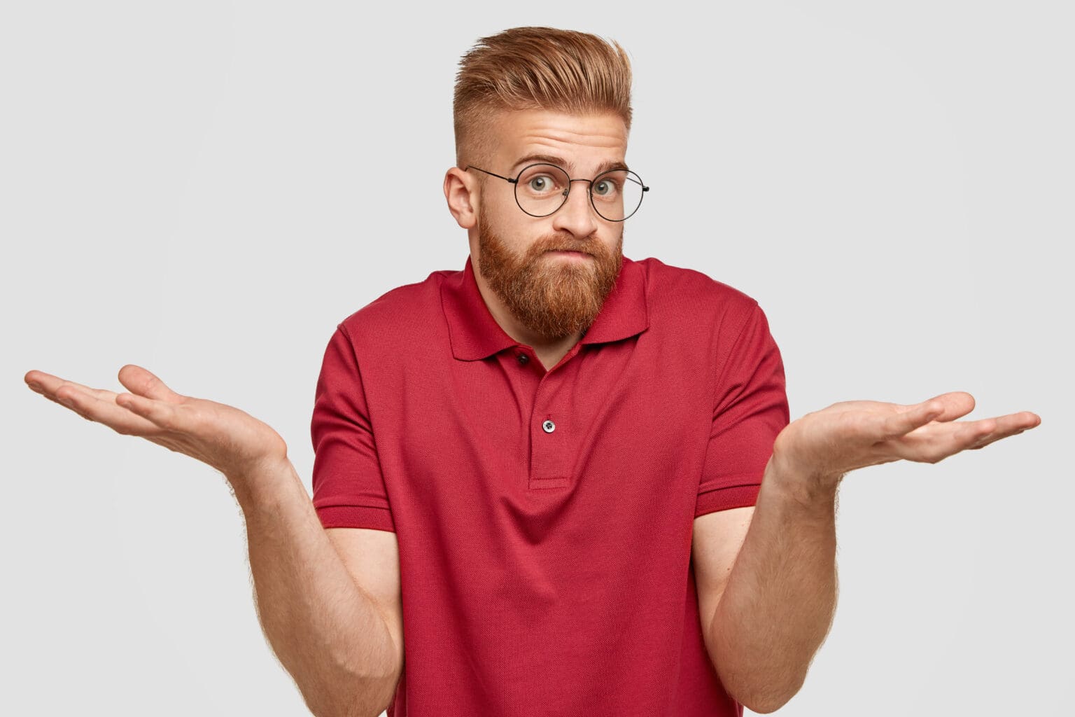 I know nothing. Handsome stylish hipster with thick ginger beard, spreads palms with questionned expression, being unsure, wears transparent glasses, poses against white background. Doubt concept